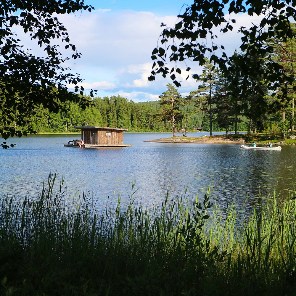 Ett trähus flyter på en lugn sjö omgiven av gröna träd, med två personer som paddlar kanot i närheten. Högt gräs ramar in fotot i förgrunden, och himlen är delvis molnig.