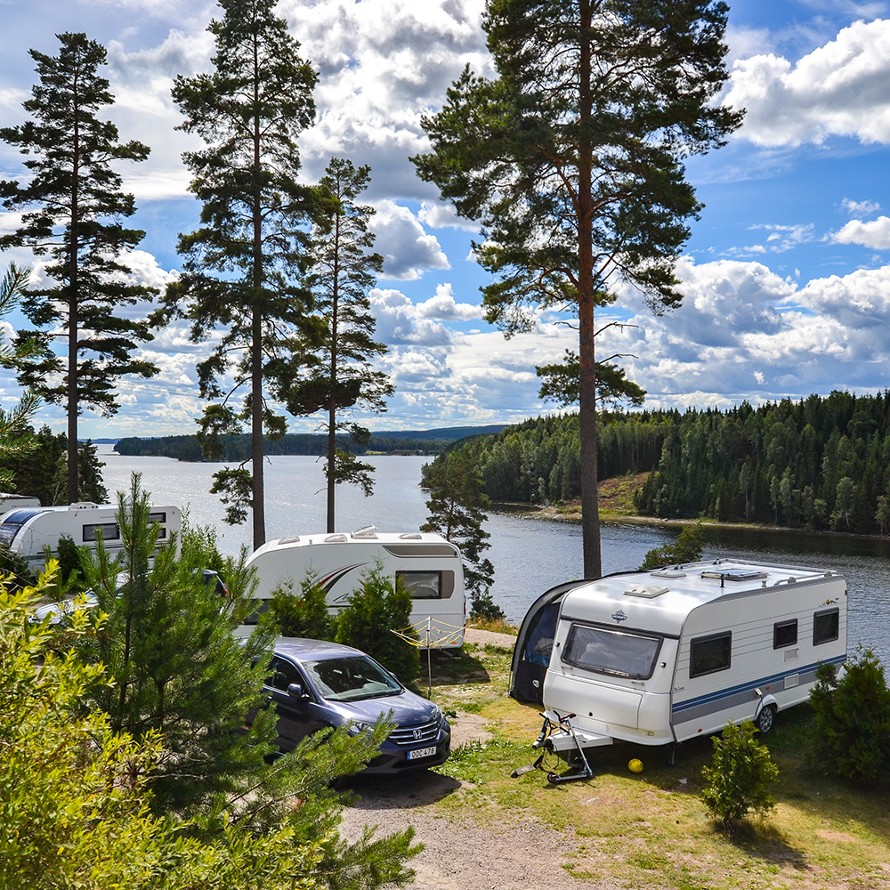 Flera husvagnar och en bil står parkerade nära en sjö, omgiven av höga tallar och frodig grönska under en delvis molnig himmel. Det lugna vattnet och de skogsklädda kullarna skapar en fridfull campingmiljö.