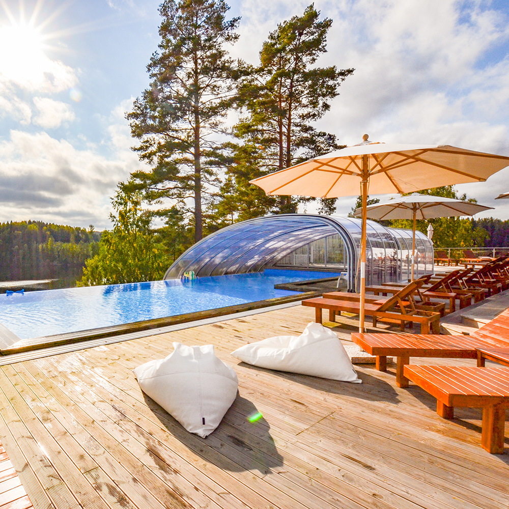 Terrass med trästolar, parasoller och vita säckar bredvid en infinitypool med utsikt över en skog och sjöar. Solen skiner och himlen är delvis molnig.