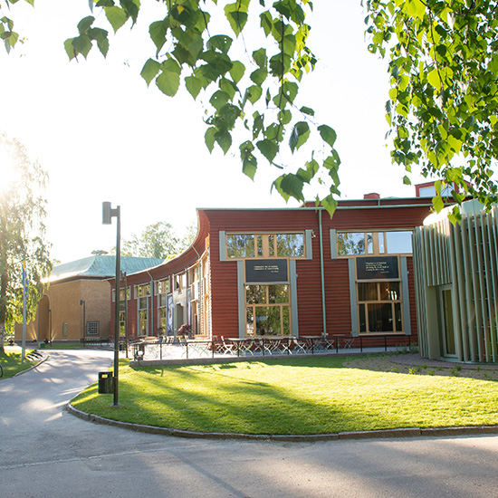 En modern röd och grön byggnad med stora fönster är omgiven av grönt gräs och träd i starkt solljus. En svängd gångväg och bänkar syns i den fridfulla utomhusmiljön.