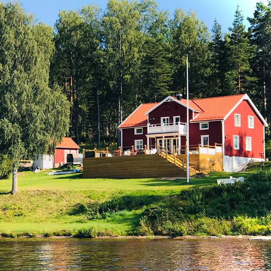 A red wooden house with white trim and a red roof stands by the water, surrounded by green grass, trees, and a wooden fence, with a forest in the background.