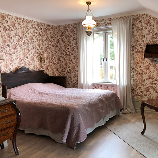 A cozy bedroom with floral wallpaper, a wooden headboard, a pink quilted bedspread, a vintage chandelier, a window with white curtains, and antique wooden furniture.