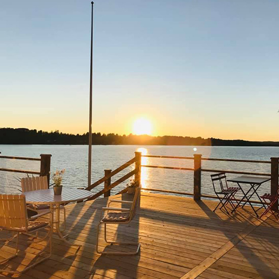 A wooden deck with white outdoor furniture overlooks a tranquil lake at sunset, with the sun low on the horizon casting long shadows. Trees line the far shore beneath a clear sky.