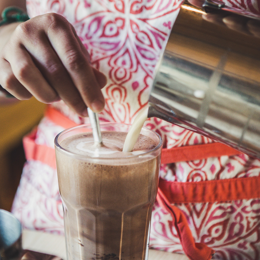 A person wearing a red-and-white-patterned apron is stirring a tall glass of iced chocolate while pouring milk into it. The drink is frothy, and the person is holding a straw.