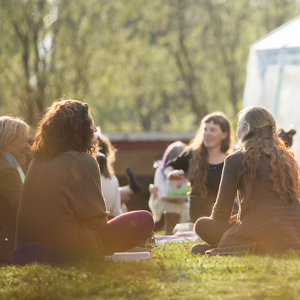 A group of women are sitting on the grass outdoors, chatting and smiling in the sunlight, with trees and a bright background behind them, creating a warm and relaxed atmosphere.