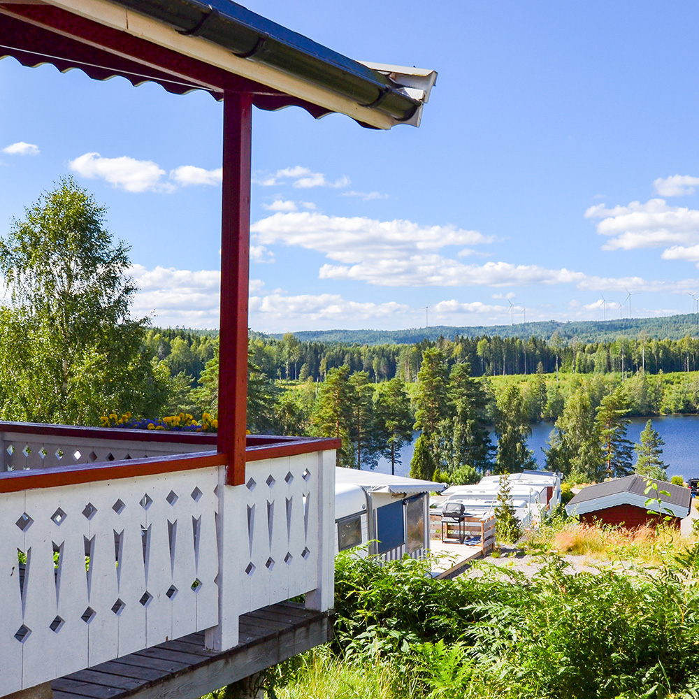 En solig balkong med ett vitt räcke har utsikt över ett naturskönt landskap med träd, en sjö, hustak och böljande kullar under en klarblå himmel med spridda moln.