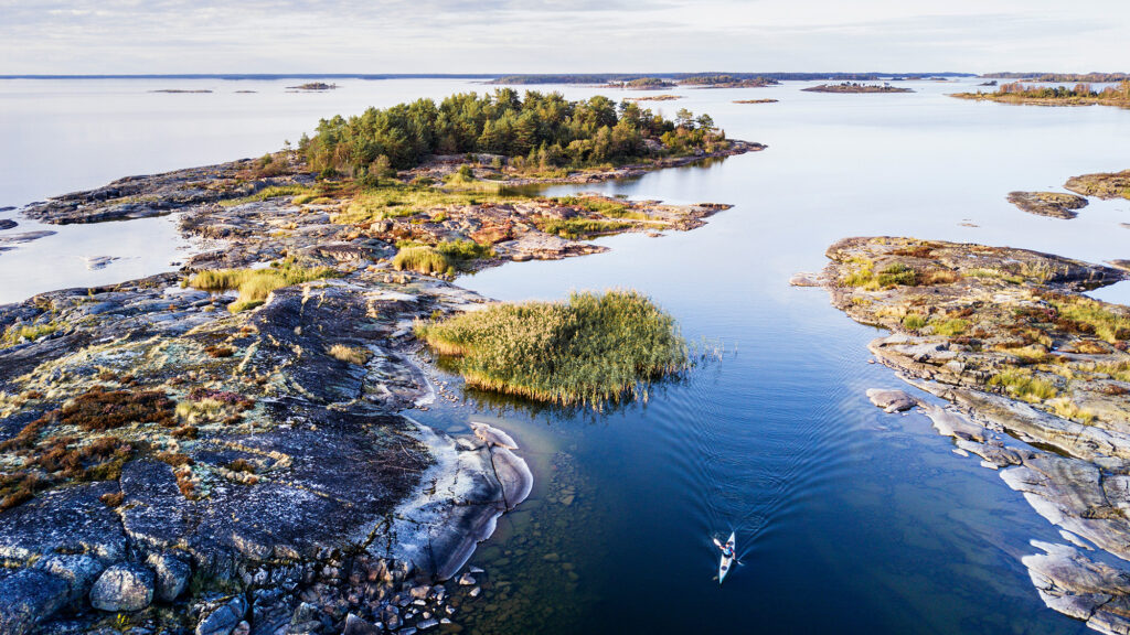 A person is kayaking through calm blue waters surrounded by rocky islands and lush greenery, with a vast sky and more small islands visible in the distance.
